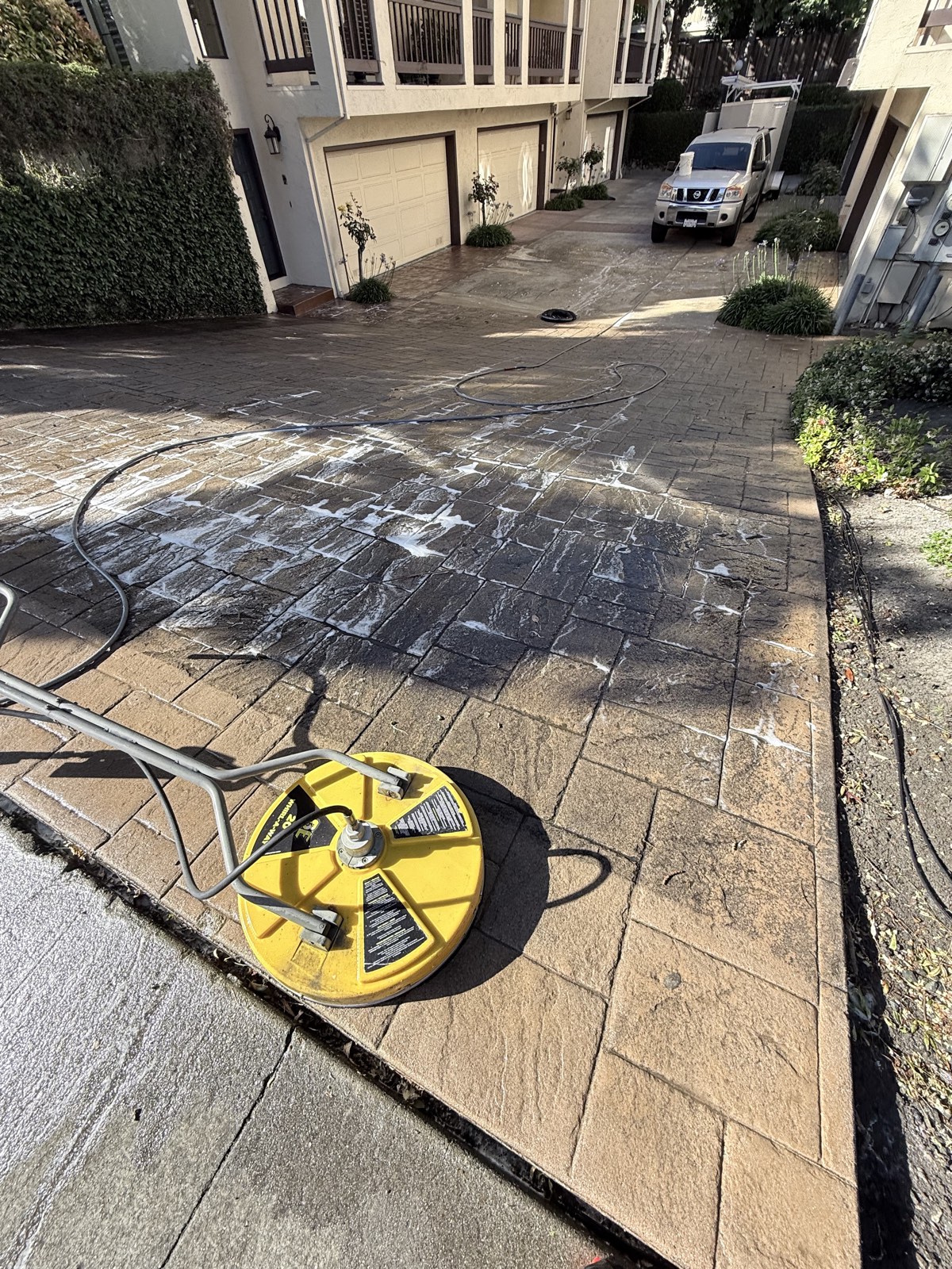 Before — dark algae-stained drive court mid-cleaning between townhome garages, detergent foam on the stamped concrete